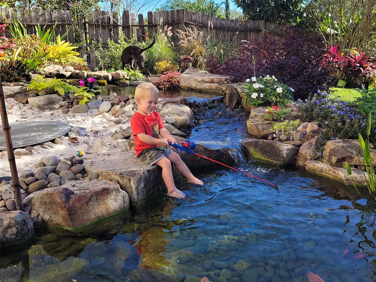 Child enjoying a fishing game beside a landscaped backyard pond with vibrant flowers and clear water.