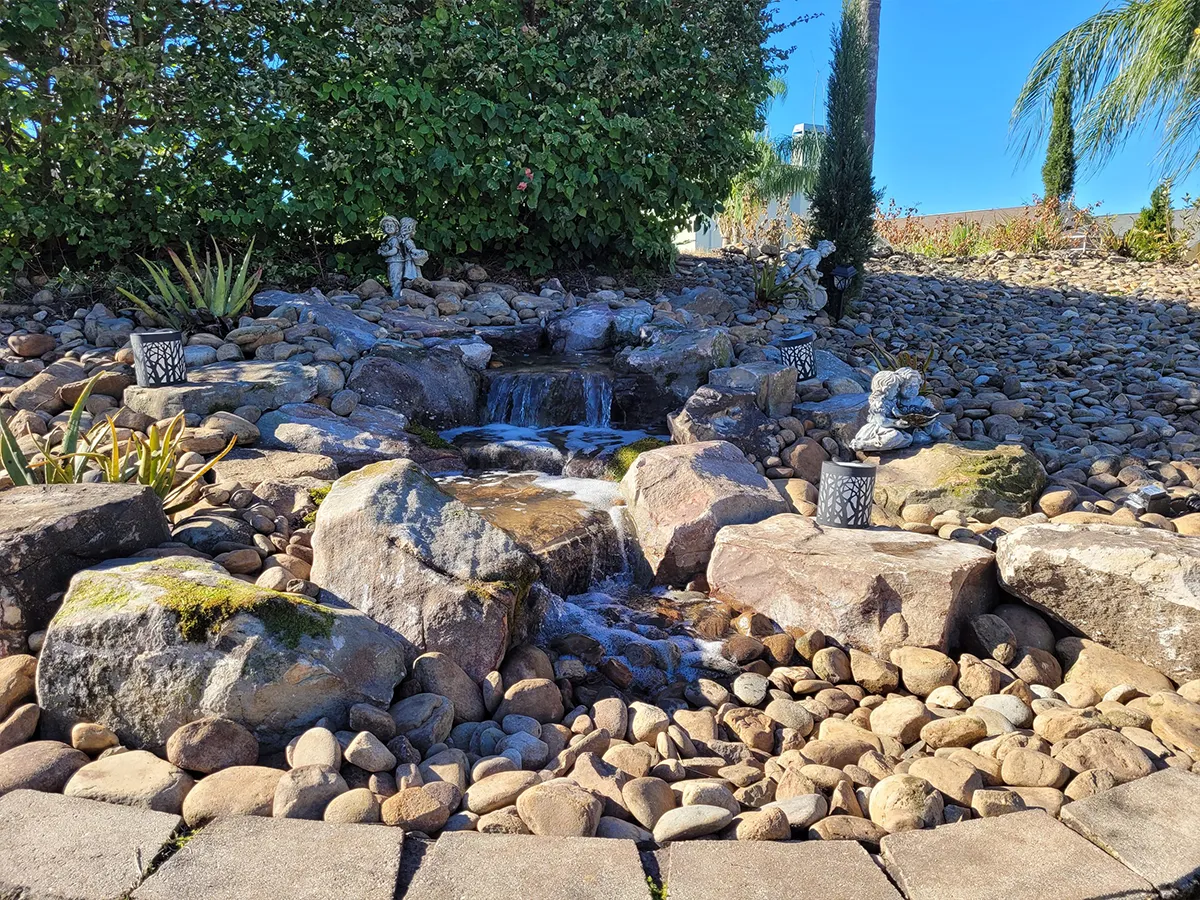 Small backyard waterfall feature with rocks, statues, and surrounding garden decor.