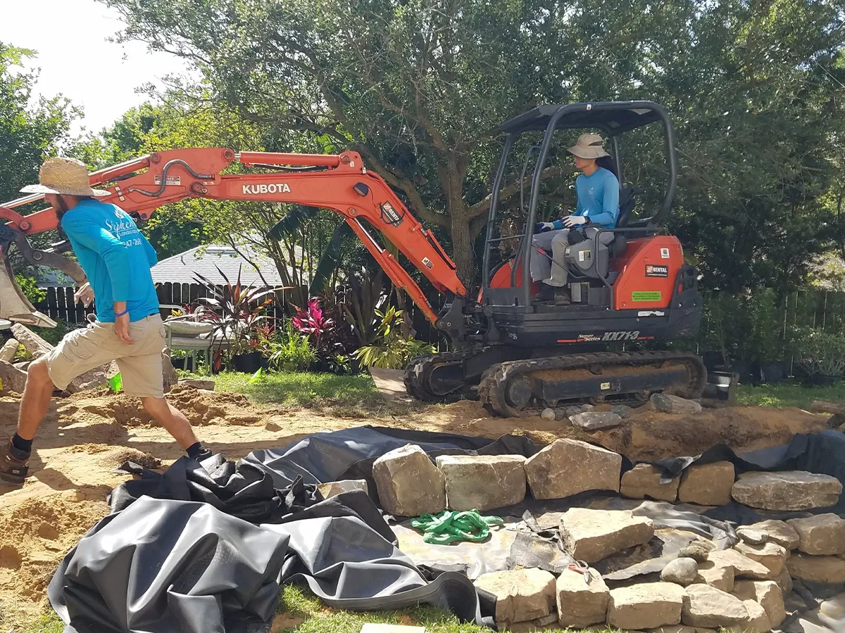 Two workers building a pond, using a small excavator and rocks, with protective lining laid out, in a garden setting with trees and plants in the background.