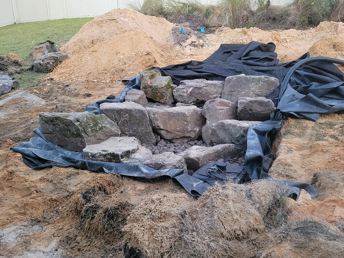 Foundation setup for a backyard pondless waterfall, featuring rocks and liner materials.