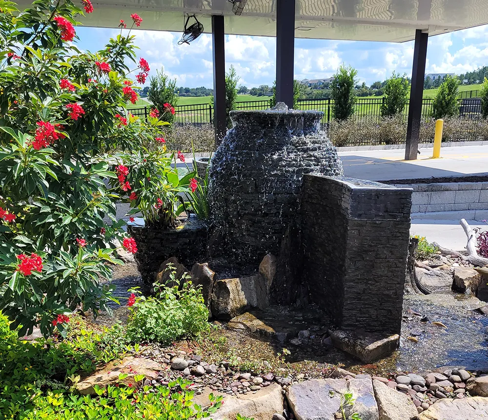 Garden fountain with cascading water over stone feature surrounded by blooming red flowers and greenery