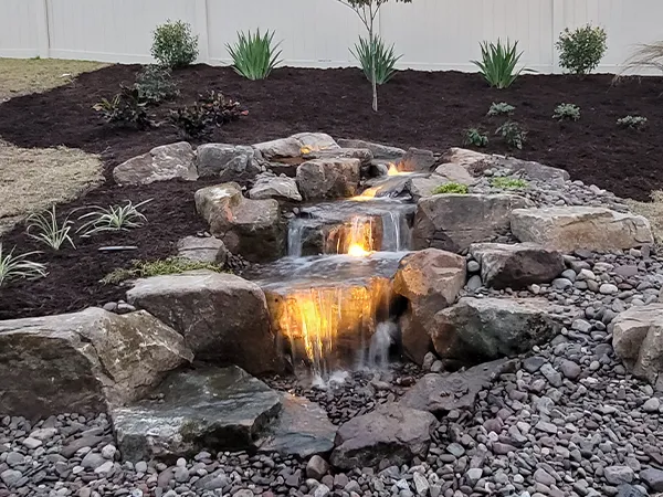 Illuminated backyard waterfall with natural stone and lush landscaping.