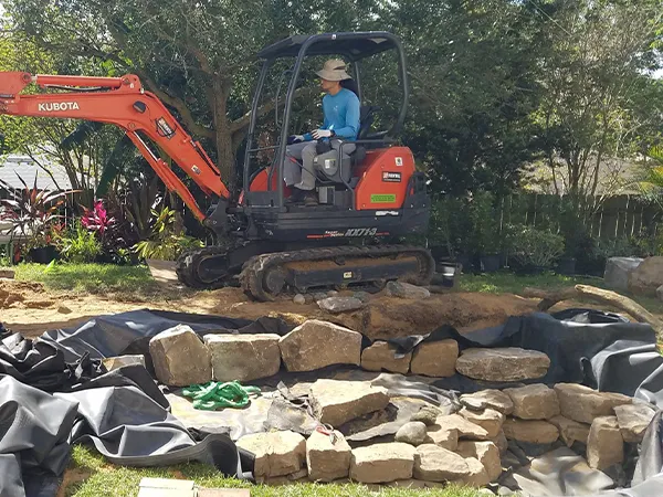 Worker operating an excavator to build a backyard pond, placing large rocks and lining to create a custom water feature.