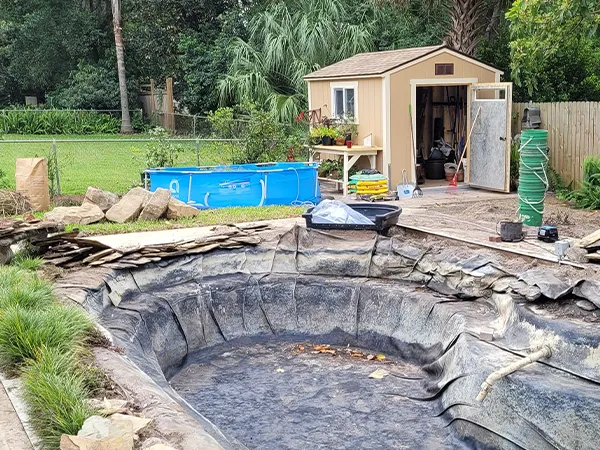 Backyard pond under repair, with large stones and landscaping fabric, preparing for renovation and pond restoration.