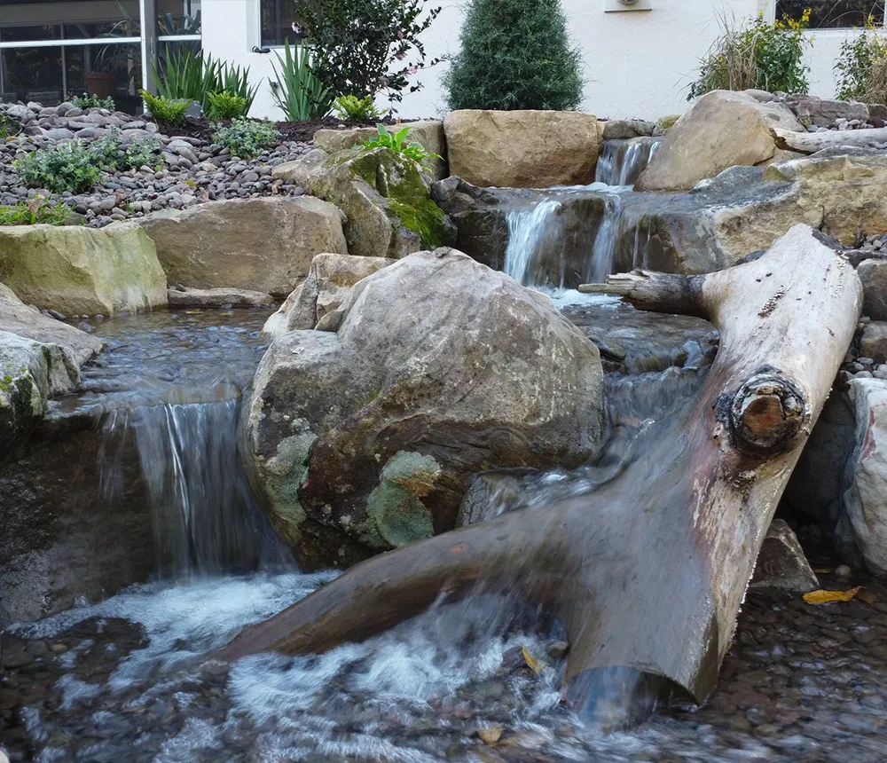 Backyard waterfall feature with natural rocks and log accents, surrounded by lush garden plants.