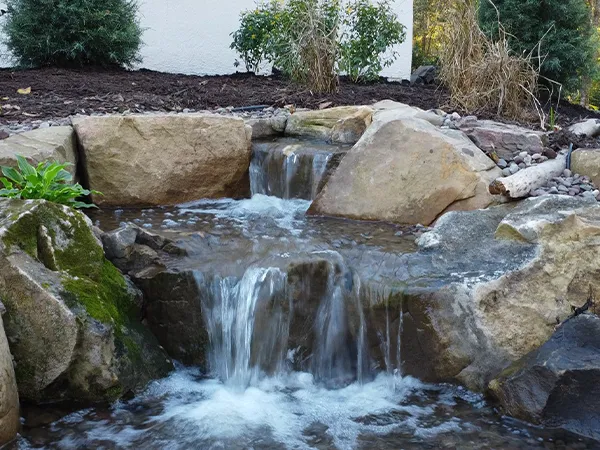 Small backyard waterfall with cascading water over natural stones, enhancing a garden landscape near a house.