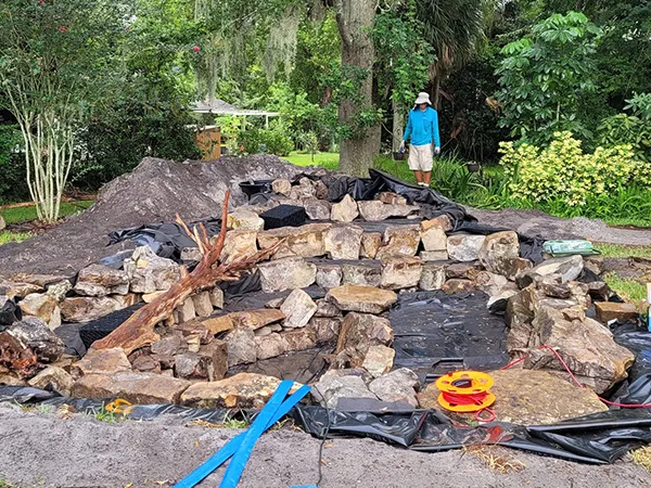 Backyard pond construction in progress with rock placement, liner installation, and landscaping work."