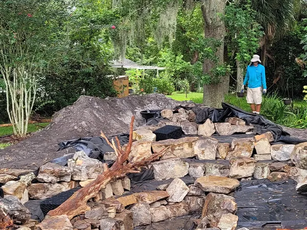 Garden pond under construction with layered rocks, a wooden log, and a person overseeing the progress in a wooded backyard.