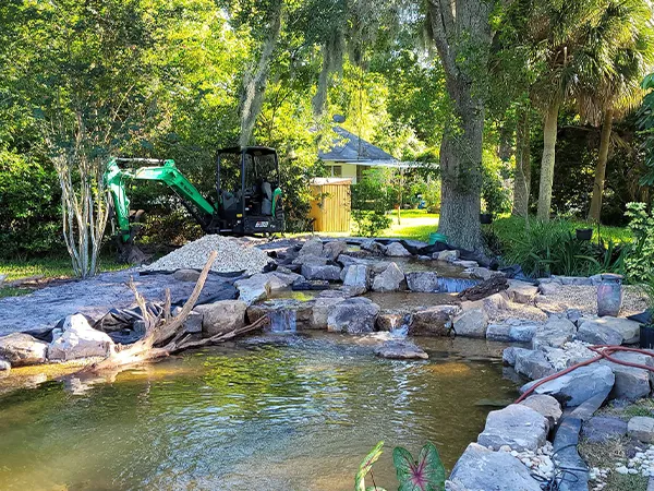 Excavator working on backyard pond installation with natural stone and waterfall features