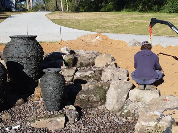 Technician setting up basalt stone pots and rocks for an outdoor water fountain installation near a driveway.